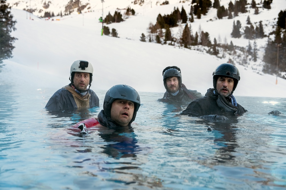 Four men wearing skiing helmets and gear are sitting in a pool, snowy mountains behind them and looking into the camera bewildered 