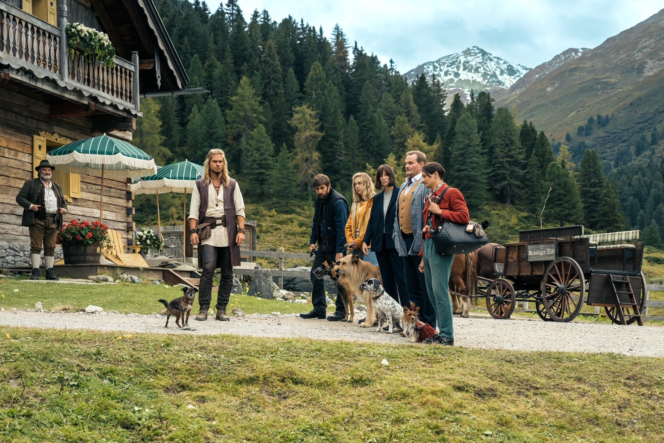 A Group of people are standing in front of a men, dressed like a modern viking. Each Person has a dog next to them,  behind them are high tyrolean mountains and a cozy looking mountain cottage