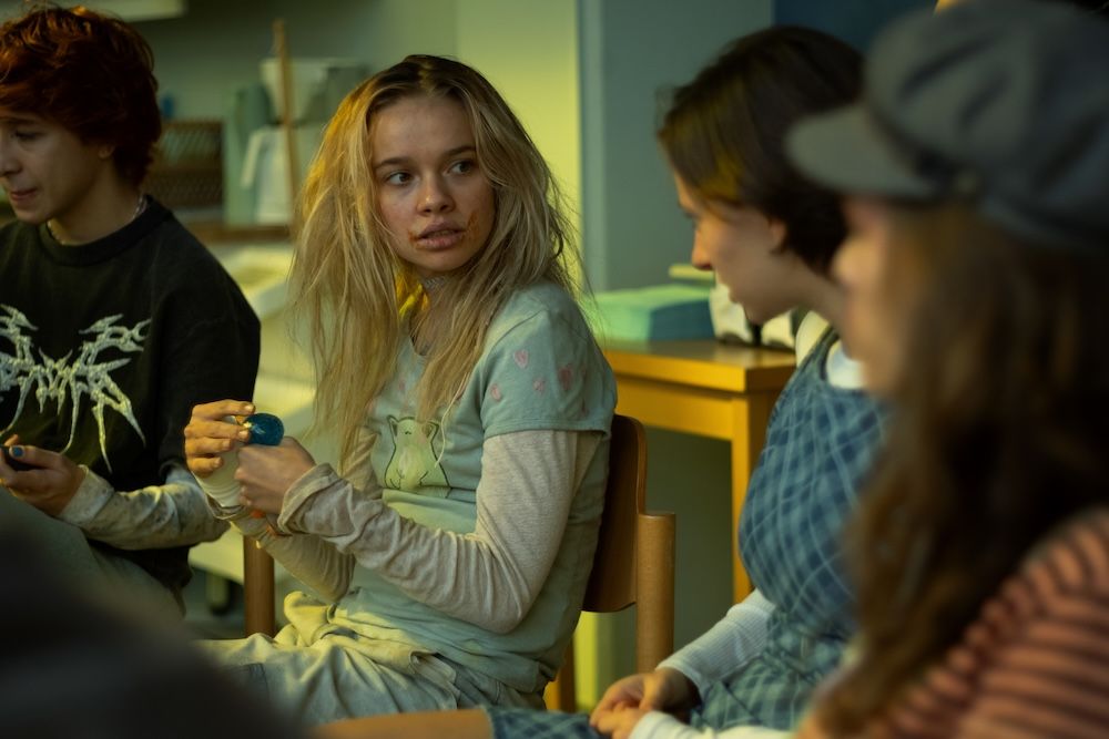A girl with messy hair and blisters around her mouth is staring shocked at a girl sitting next to her in a group therapy session