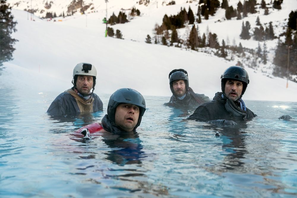 Four men wearing skiing helmets and gear are sitting in a pool, snowy mountains behind them and looking into the camera bewildered