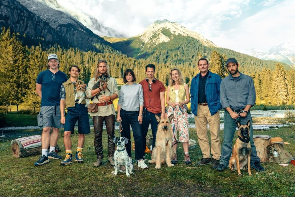 A Group of eight people stand next to each other, looking into the camera, Tyrolean mountains behind them and five dogs of different breeds in front of them