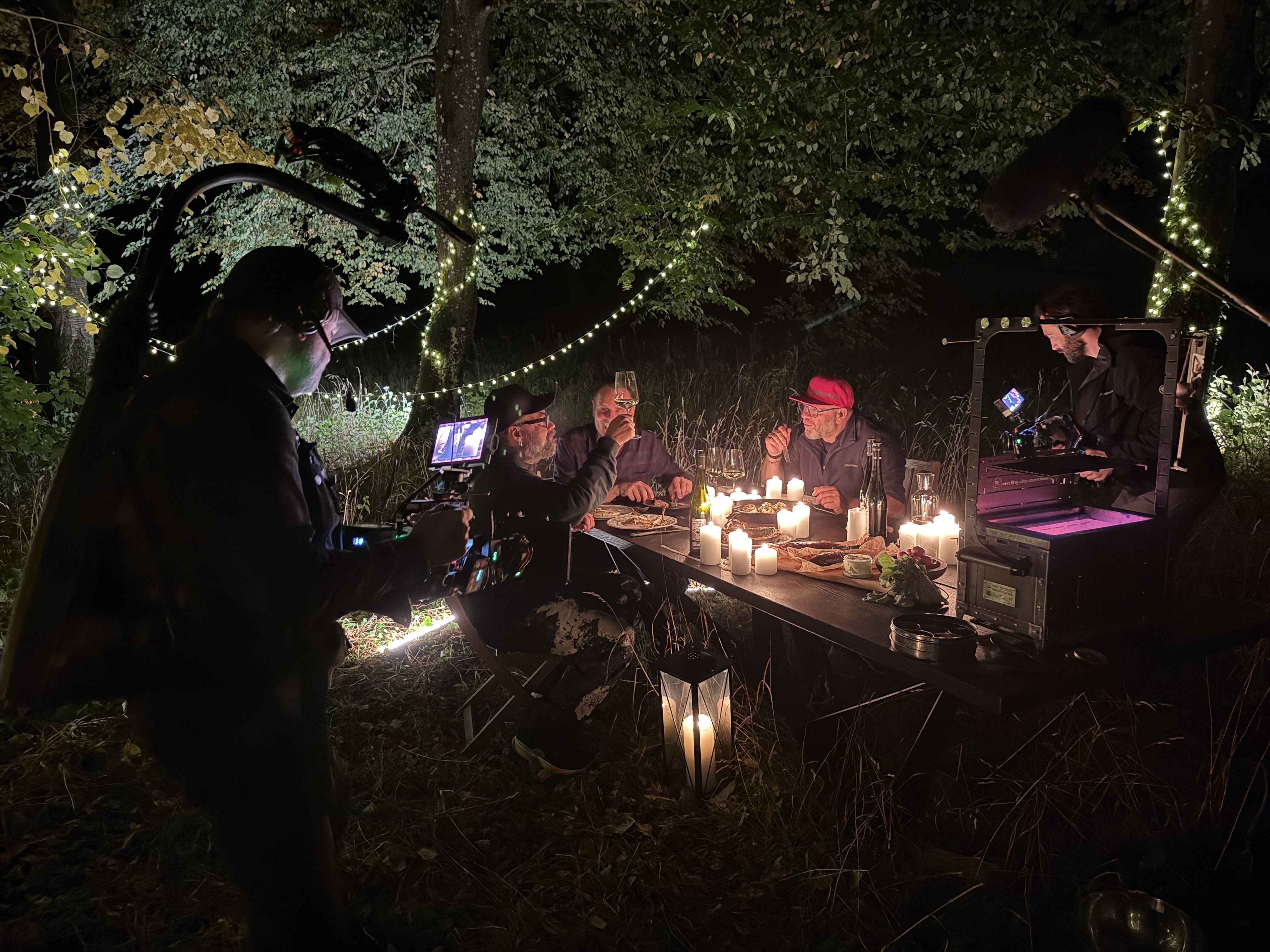 Three man are sitting by a table, it is dark they are in nature surrounded by fairy lights and trees. They are eating, one is raising a glass of wine. It is a behind the scenes photo, the camera operators can be seen in the photo.
